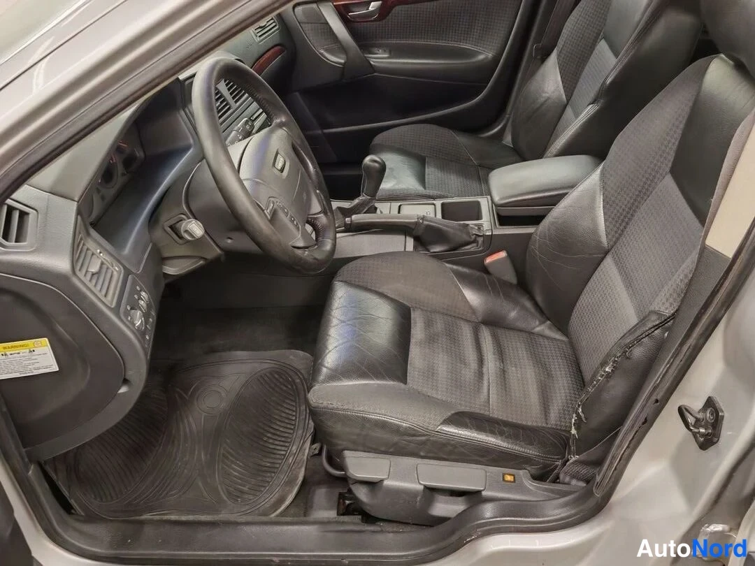 Muu: Volvo V70:n etummat istuimet ja kojelauta tummilla verhoilla, ohjauspyörä ja manuaalivaihteen kahva näkyvät.