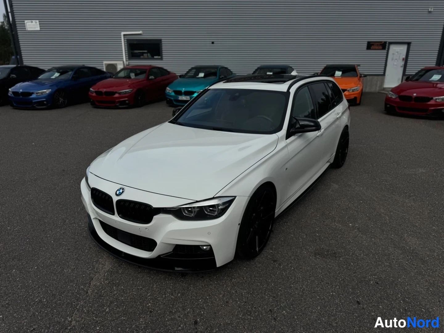 Etuviisto, nokka vasemmalle: BMW 3-sarjan farmari valkoisena etunurkkakuvassa, mustat vanteet ja takana useita muita BMW-autoja.