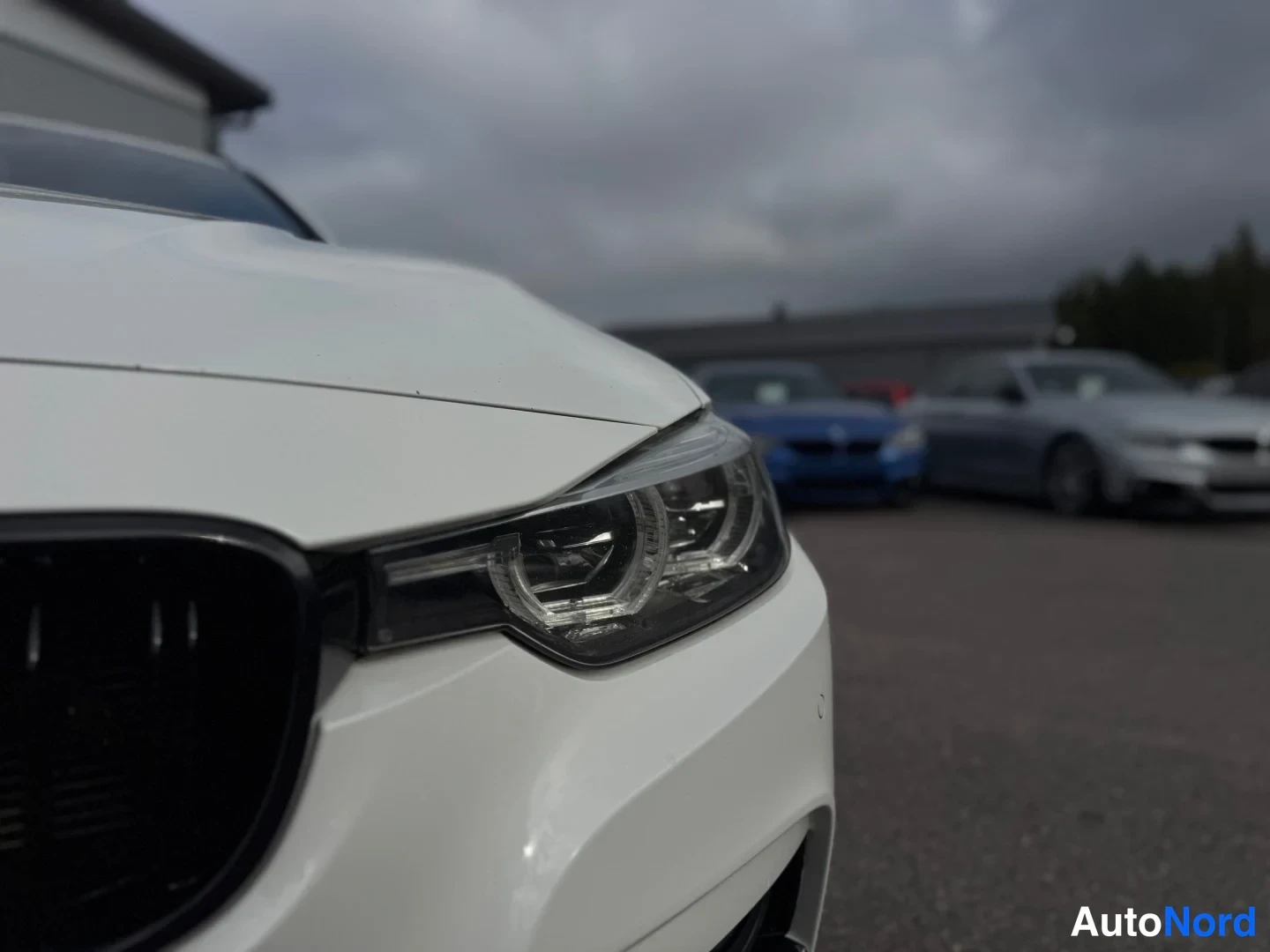 Etuviisto, nokka vasemmalle: Close-up of the front-right corner of a white BMW showing part of the grille, headlight, and bumper with background cars on asphalt under cloudy sky.