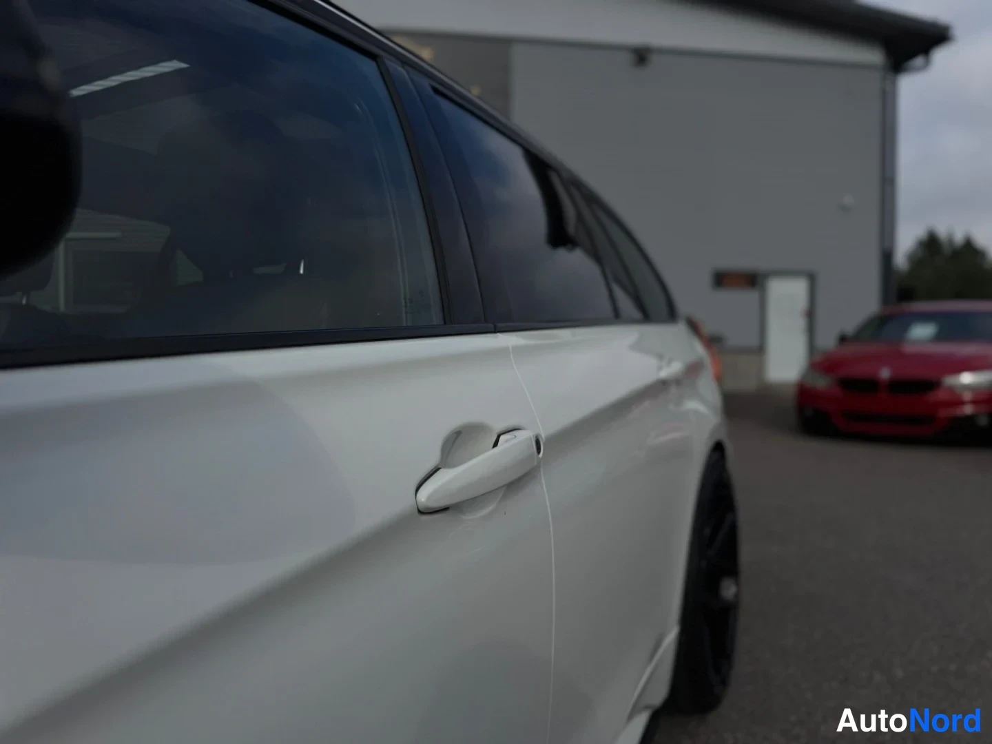 Sivulta: Sivukuva valkoisen auton takaovesta ja mustasta renkaasta, taustalla punainen auto ja harmaa rakennus.