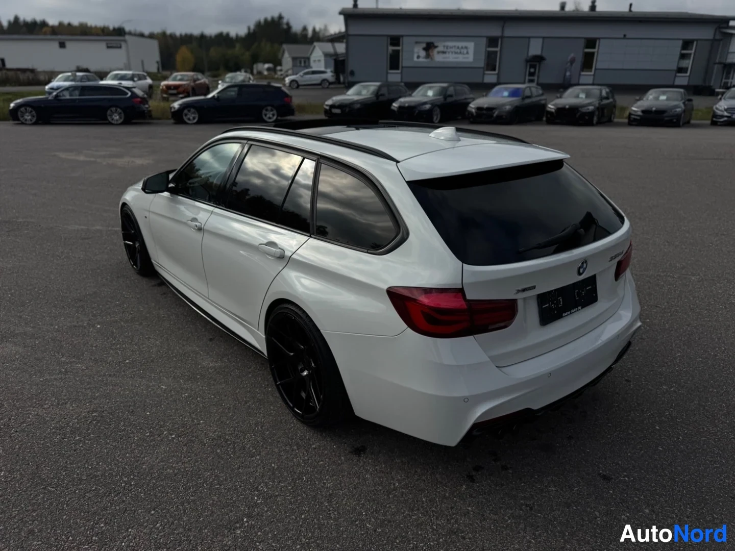 Takaviisto: Valkoinen farmariauto takaviistosta vasemmalle kuvattuna; takaosa ja vasen kylki näkyvät, mustat vanteet ja tummat lasit.