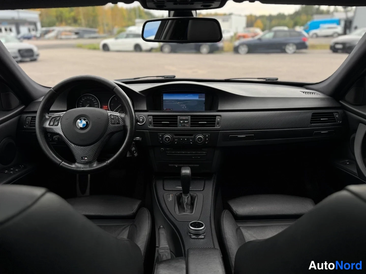 Etuviisto: Kojelauta ja ohjauspyörä takaa päin kuvattuna; kosketusnäytöllinen navigaatio, automaattivaihteen valitsin ja BMW-ohjauspyörä näkyvät.