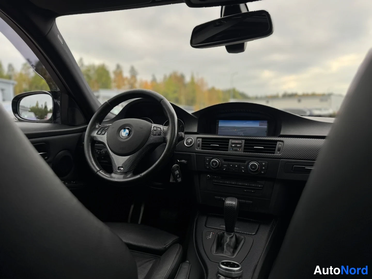 Muu: Kuljettajan puolen sisätila: BMW-ohjauspyörä, tumma kojelauta navigointinäytöllä, automaattivaihteen valitsin ja sivupeili.