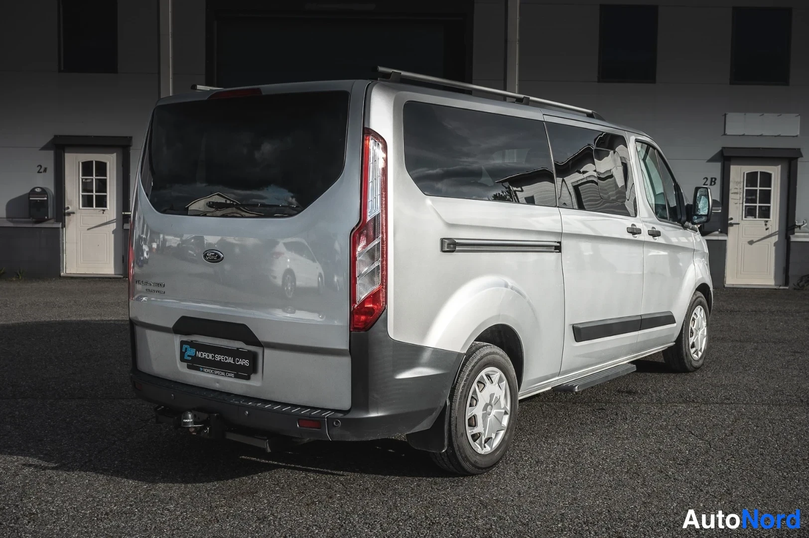 Takaviisto: Hopeanvärinen Ford Tourneo Custom -pakettiauto taka-vinoottikuvassa, perävaunukoukku ja kattotelineet näkyvissä.