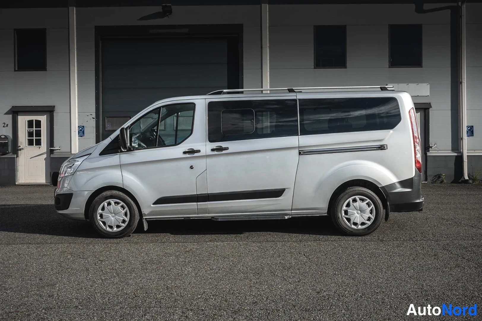 Sivulta: Hopeanvärinen Ford Tourneo Custom -pakettiauto sivukuvassa vasemmalta, kattotelineet ja tummat ikkunat näkyvissä.