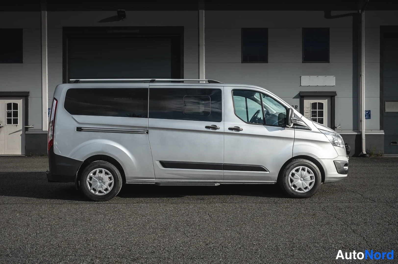 Etuviisto, nokka oikealle: Hopeanvärinen Ford Tourneo Custom -pakettiauto sivukuvassa oikealta, kattotelineet ja etusuunnan valot osittain näkyvissä.