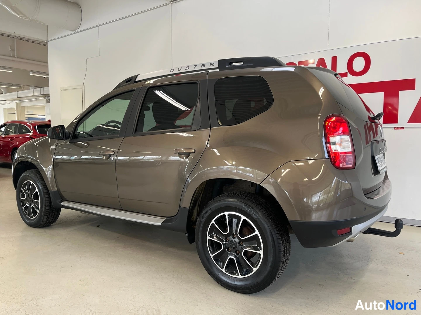 Takaviisto: Dacia Duster -maasturi takaviistosta kuvattuna sisätiloissa, ruskeakorinen, takaosa ja kylki näkyvissä, kattokaiteet ja vetokoukku näkyvissä.