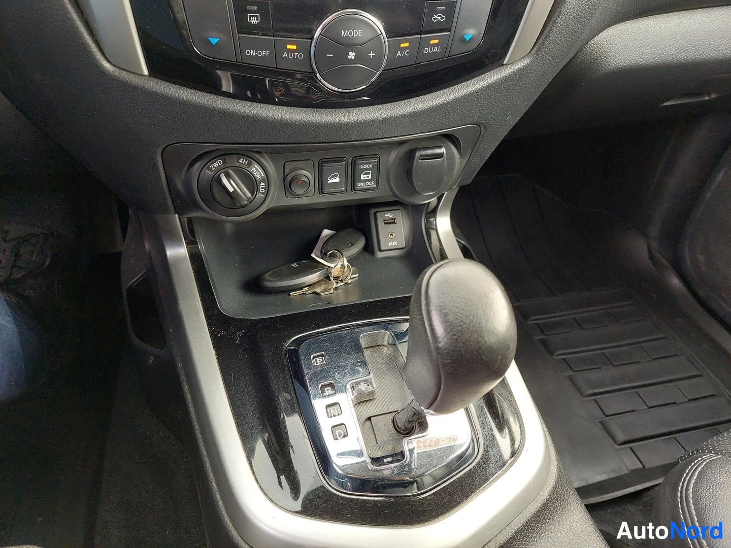 Yksityiskohta: Keskikonsolin näkymä: automaattivaihteen vaihtokeppi, ilmastointi- ja ajotilakytkimet (2WD, 4H, 4LO), USB- ja AUX-liittimet sekä avainringsi säilytyslokerossa.