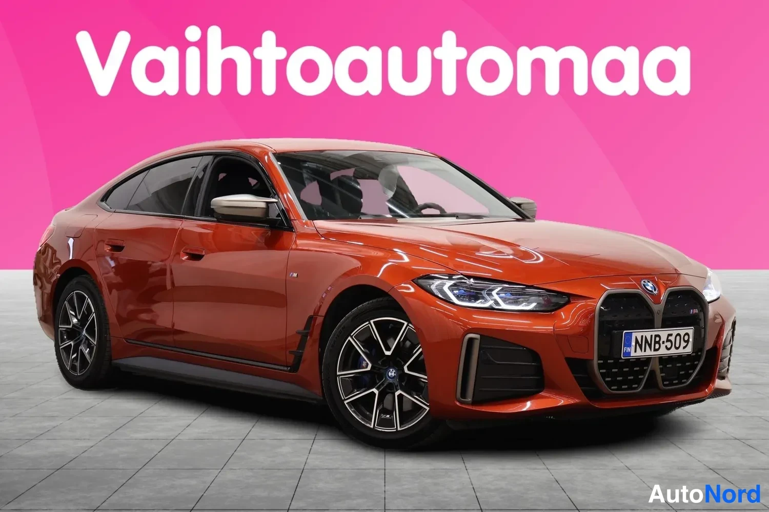 Etuviisto, nokka oikealle: BMW i4 M50 Gran Coupé kolmivarten etu- ja sivunäkymässä, etuosa oikealla; oranssinpunainen koripinta, musta etugrilli ja rekisterikilpi