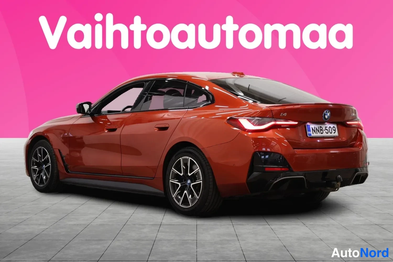 Takaviisto: Punaisenoranssi BMW i4 -henkilöauto takaviistosta kuvattuna, rekisterikilpi, takana näkyy vetokytkin.