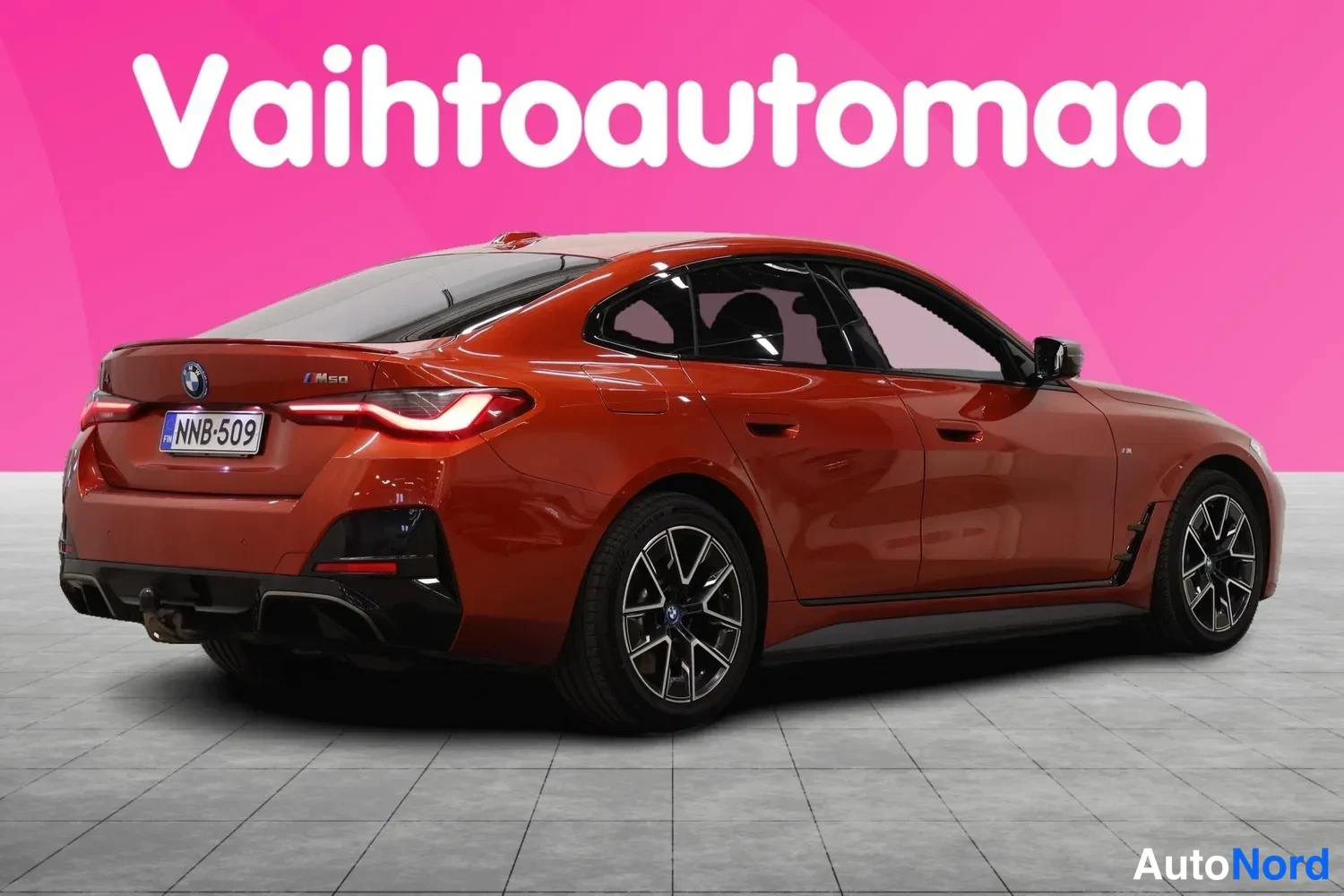 Takaviisto: Punaisenoranssi BMW i4 -henkilöauto toisesta takaviistosta kuvattuna, rekisterikilpi ja M50-merkintä takana.