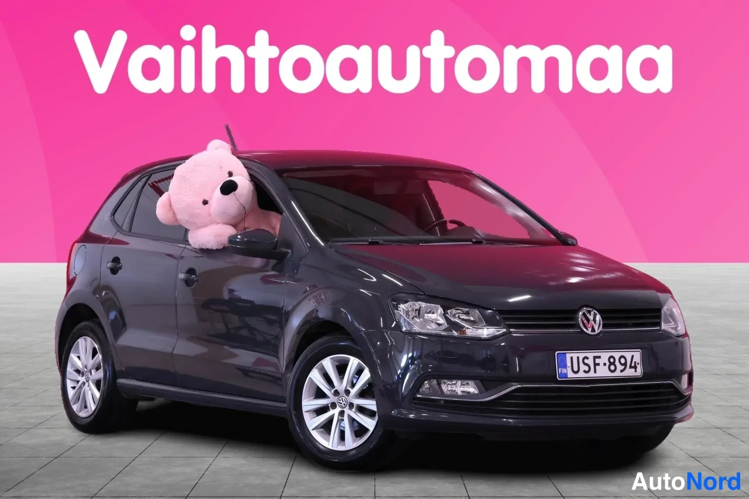 Etuviisto, nokka oikealle: Tummanharmaa Volkswagen Polo napavalokuvassa edestä oikealta vasemmalle kallistettuna, rekisteritunnus, vaaleat metallivanteet.