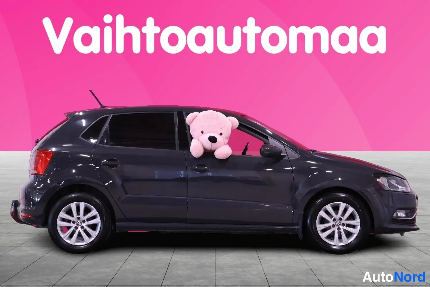 Etuviisto, nokka oikealle: Tummanharmaa Volkswagen Polo sivuprofiilista oikealta katsottuna, pehmolelu takana ikkunassa, vaaleat metallivanteet.