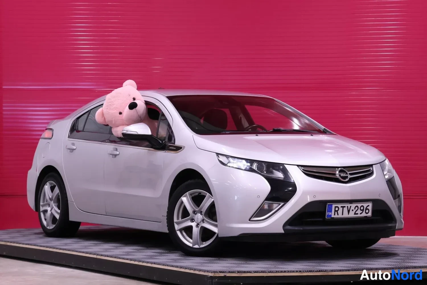 Etuviisto, nokka oikealle: Hopeanvärinen Opel Ampera etuviistosta oikealta, rekisteritunnus, pehmolelu takana oikeassa etuikkunassa, punainen tausta.