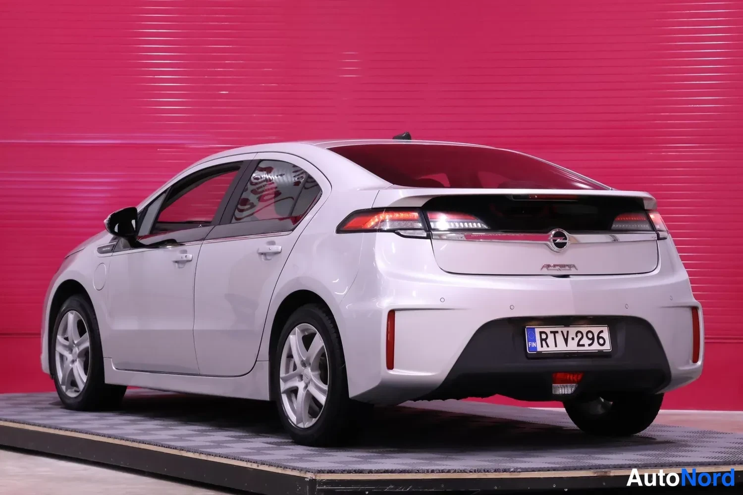 Takaviisto: Hopeanvärinen Opel Ampera takaviistosta vasemmalta, rekisteritunnus, punainen tausta.