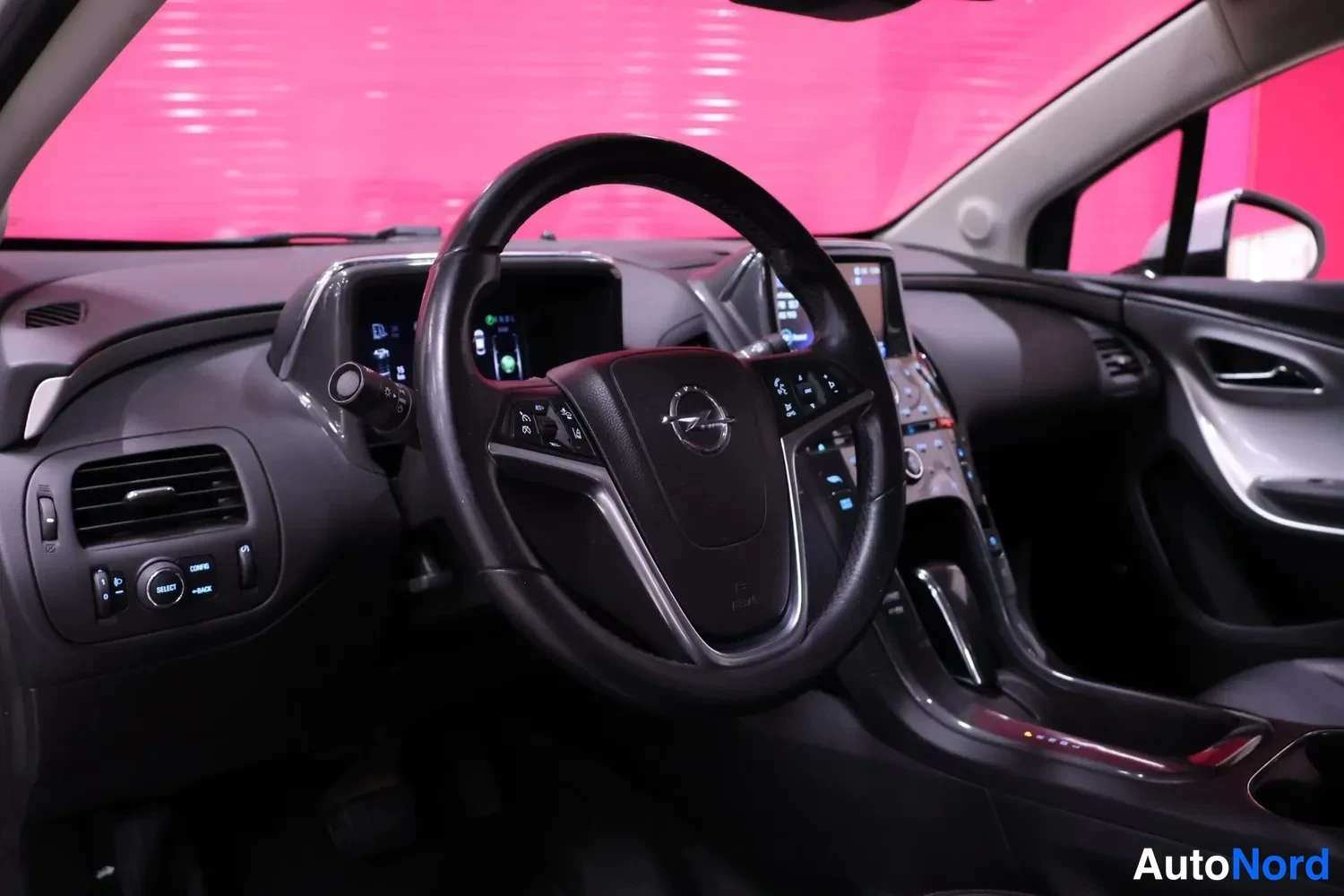 Yksityiskohta: Ohjaamo näkyy kuljettajan puolelta: Opel-merkkinen ohjauspyörä, mittariston näyttö ja keskikonsolan infotainment-näyttö ovat päällä, taustalla vaaleanpunainen s