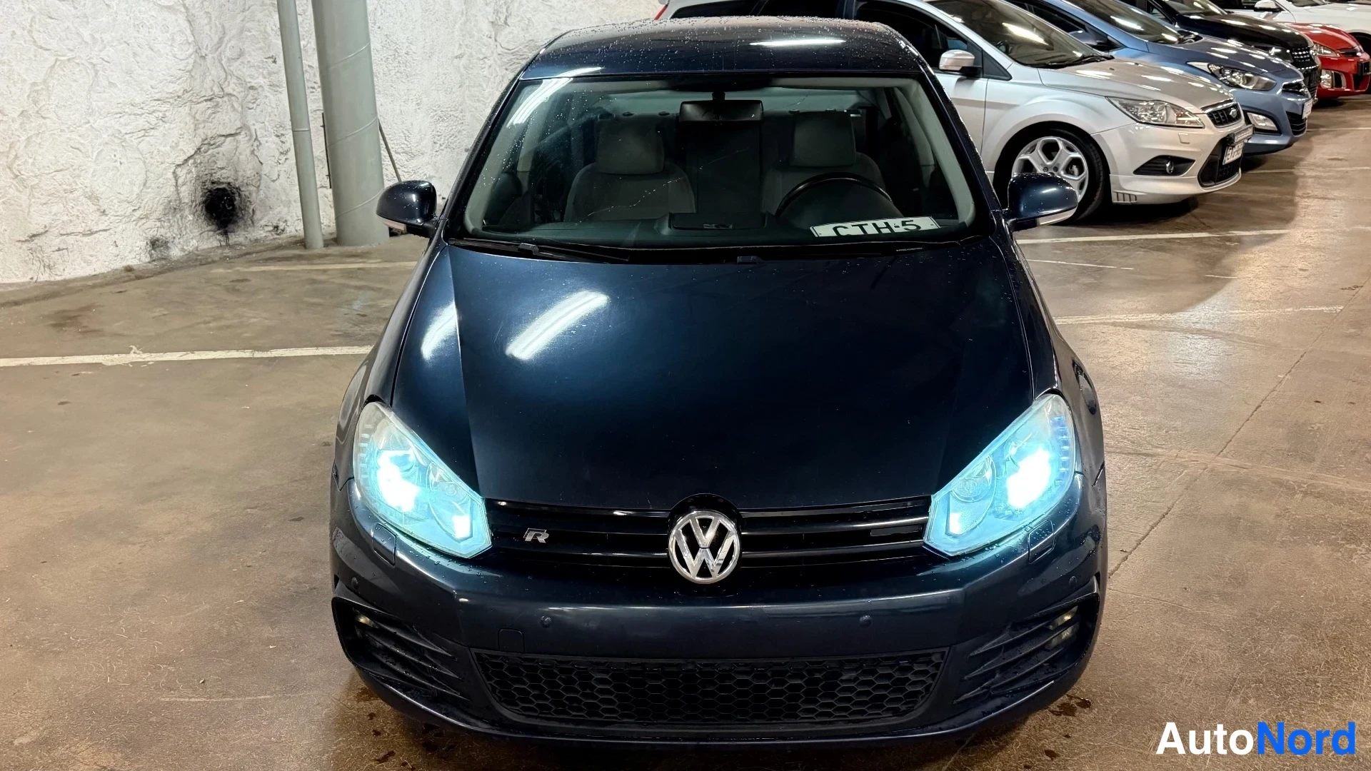 Edestä: Tummansininen Volkswagen Jetta suoraan edestä kuvattuna, syttyneet ajovalot ja R-merkki etusäleikössä.