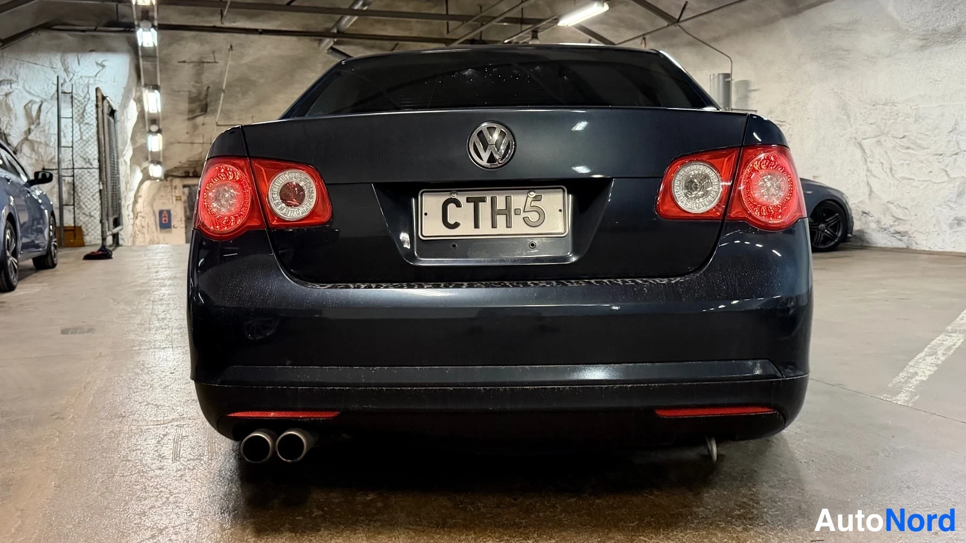Takaa: Auton takaosa suoraan takaa: tummansinisen musta Volkswagen-sedan, rekisteritunnus, kaksiosainen pakoputki.