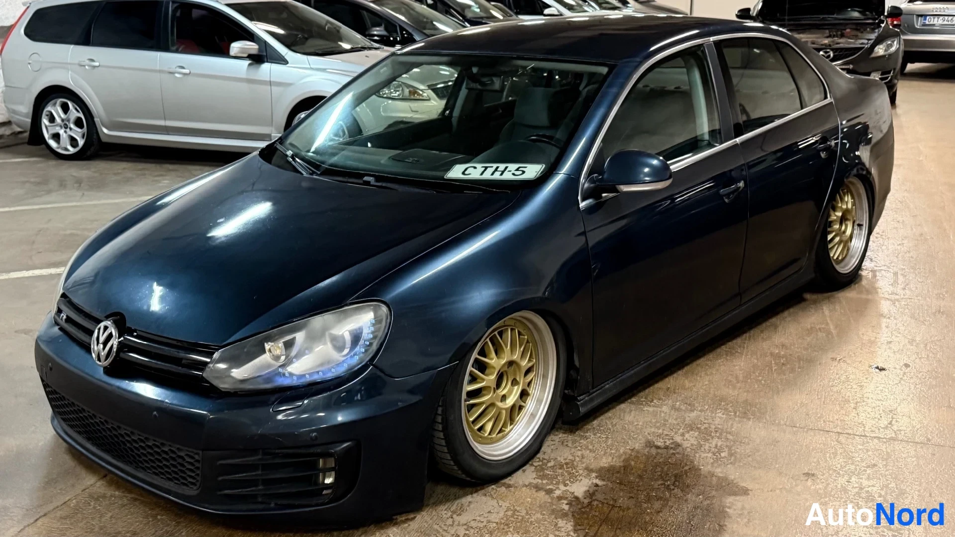 Etuviisto, nokka oikealle: Tummansininen Volkswagen Jetta -sedan ulkoa edestä oikealta kulmasta, -tarra tuulilasissa.