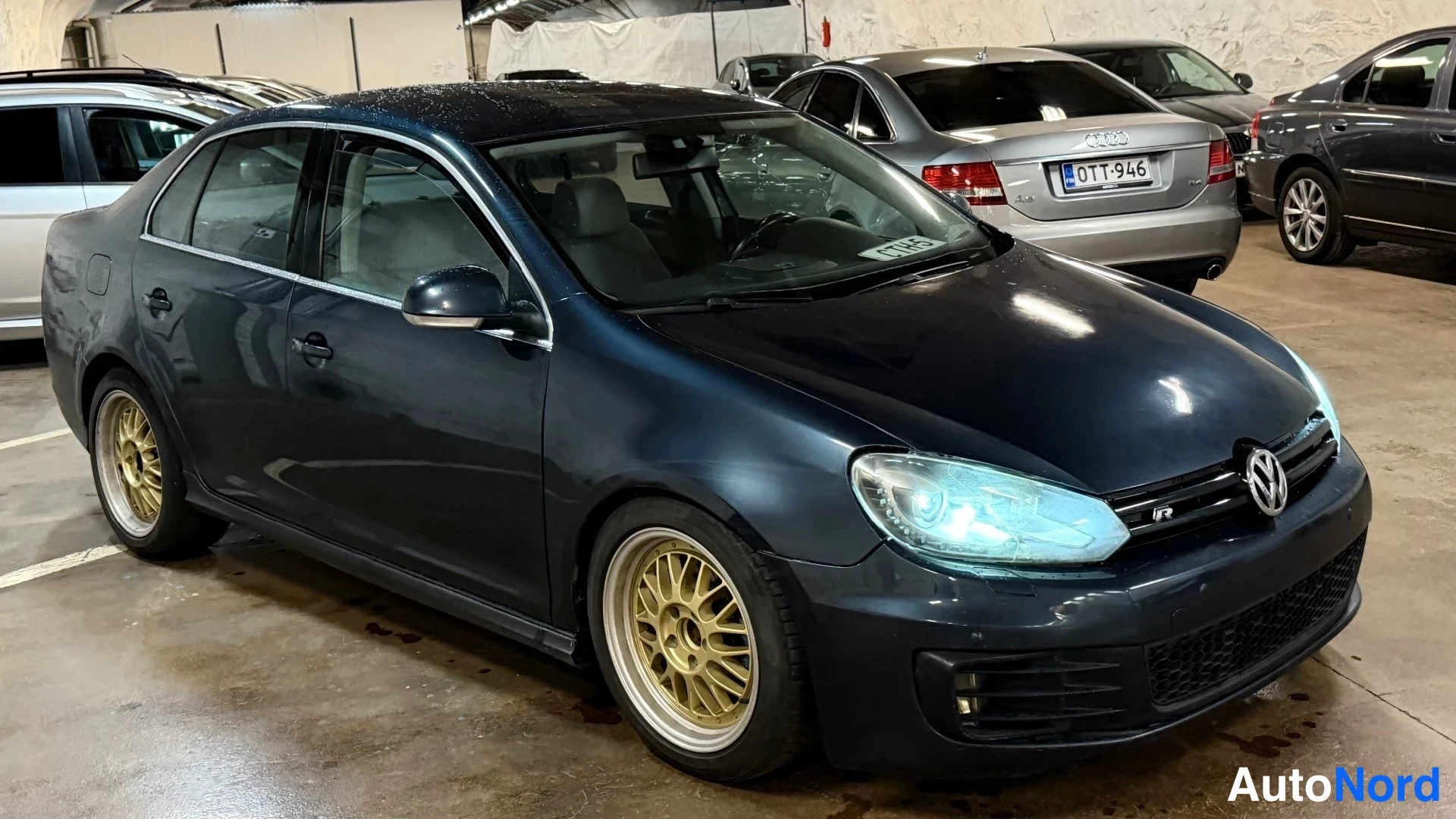 Etuviisto, nokka oikealle: Tummansininen Volkswagen Jetta -sedan ulkoa edestä oikealta kulmasta kuvattuna, kultavanteiset vanteet.
