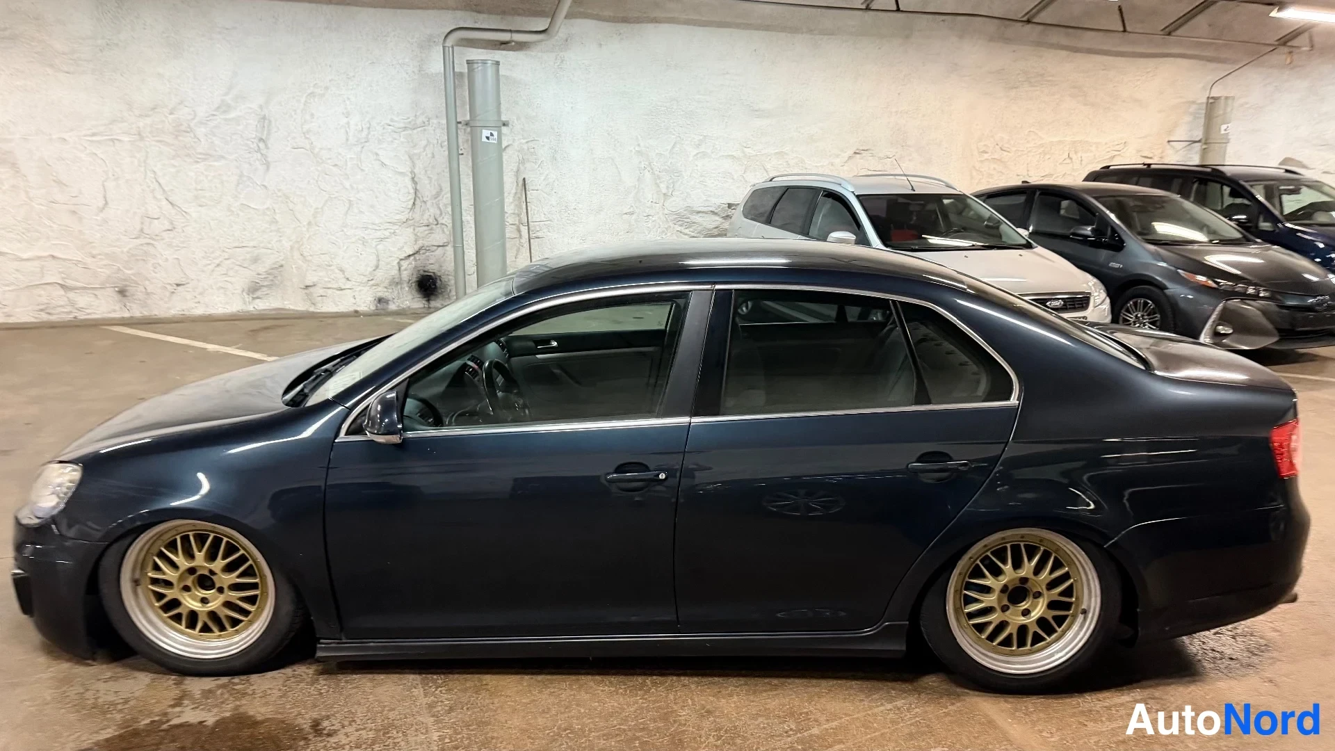 Etuviisto, nokka vasemmalle: Tummansininen Volkswagen Jetta -sedan vasemmalta sivulta etuviistoon kuvattuna, kultaväriset vanteet.