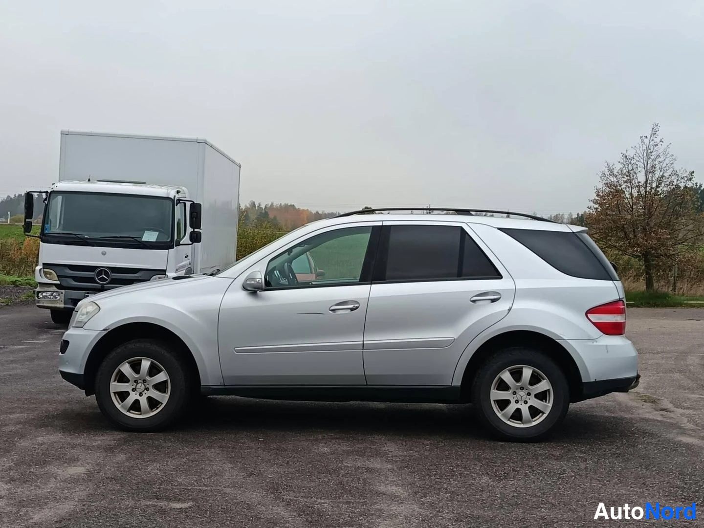 Sivulta: Hopeinen Mercedes-Benz ML -maasturi sivulta vasemmalta katsottuna parkkipaikalla kuorma-auton edessä.