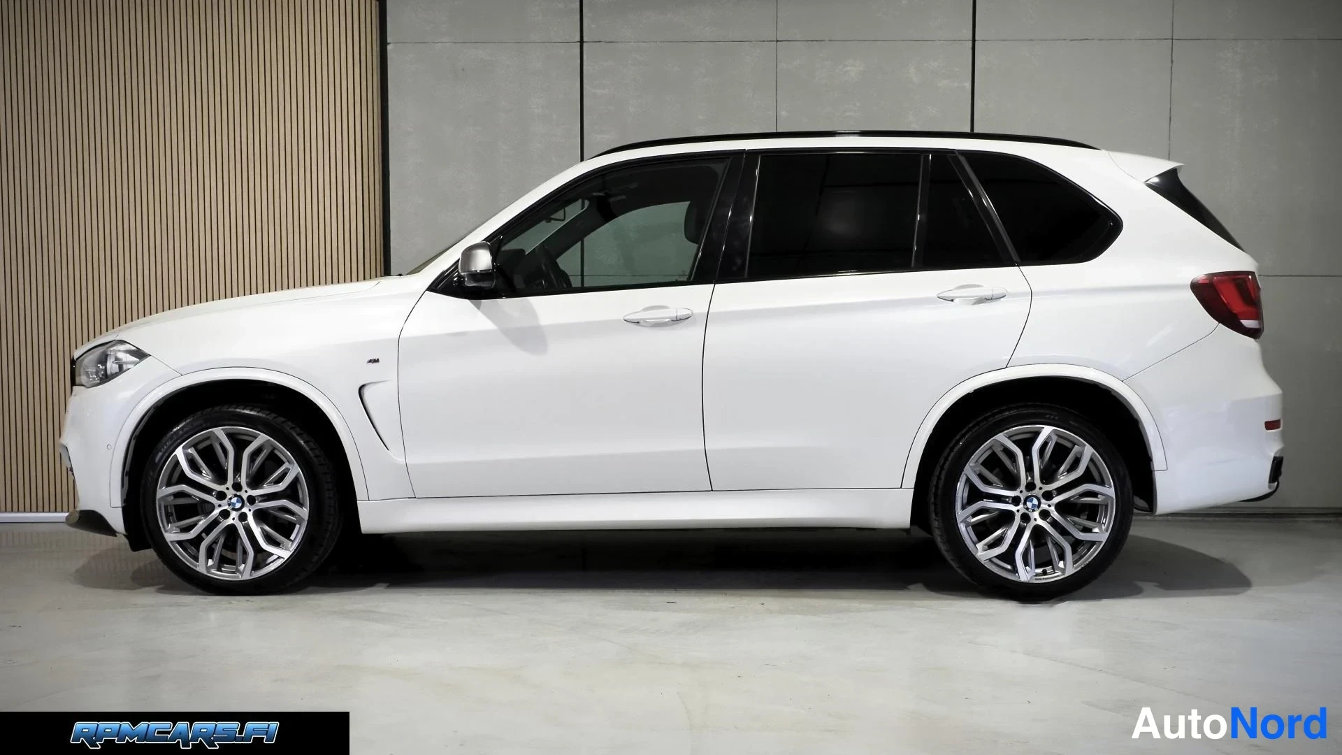 Sivulta: Valkoinen BMW X5 -maastauto sivulta kuvattuna sisätiloissa, matalapenkainen profiili ja kaksiväriset kevytmetallivanteet; takaoven lasit tummat.