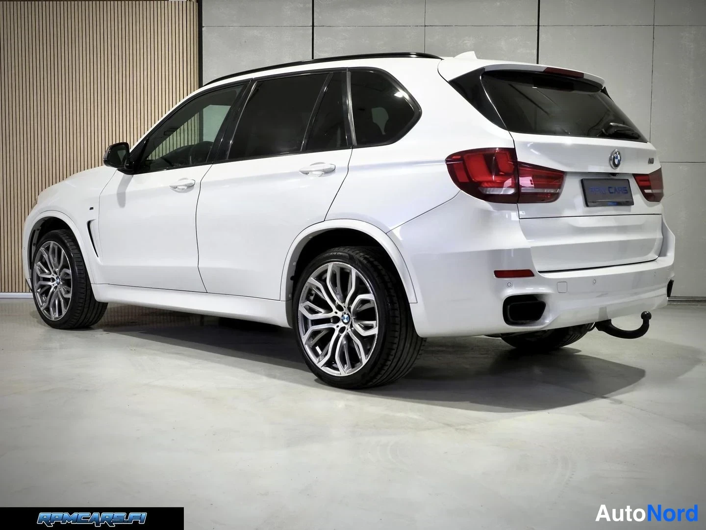 Takaviisto: Valkoinen BMW X5 takaviistosta kuvattuna sisätiloissa; takavalot, takapuskuri ja kiinnitetty vetokoulu näkyvät, takavanteet kaksiväriset.