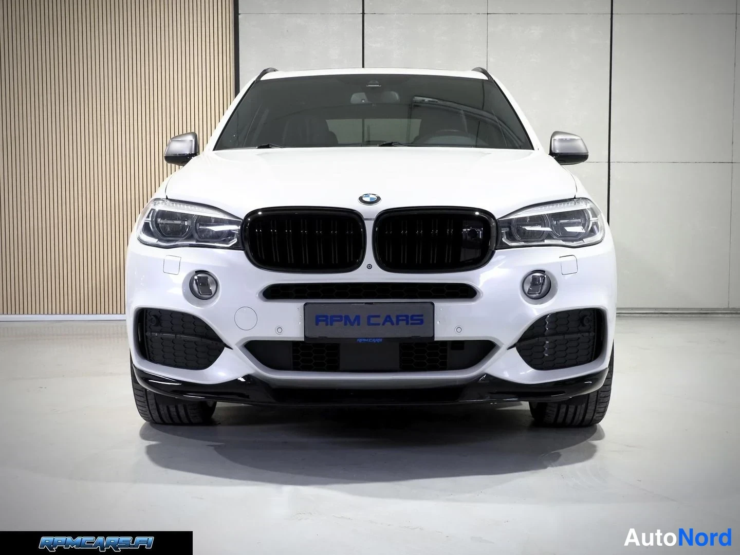 Edestä: Auton etupää suoraan edestä valokuvattuna sisätiloissa; valkoinen koripaneeli, musta etusäleikkö, BMW-merkki kiinnitetty, LED-ajovalot ja tummat puskurin ilmano