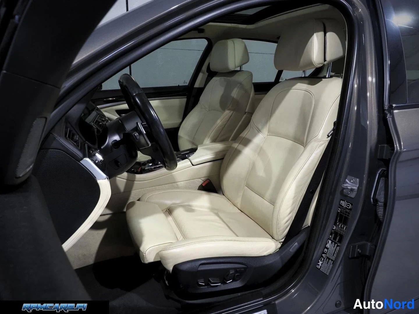 Muu: BMW:n sisätilat kuljettajan oven puolelta: beige nahkapenkki ja M-tyylinen ohjauspyörä, lasikatto yläpuolella.