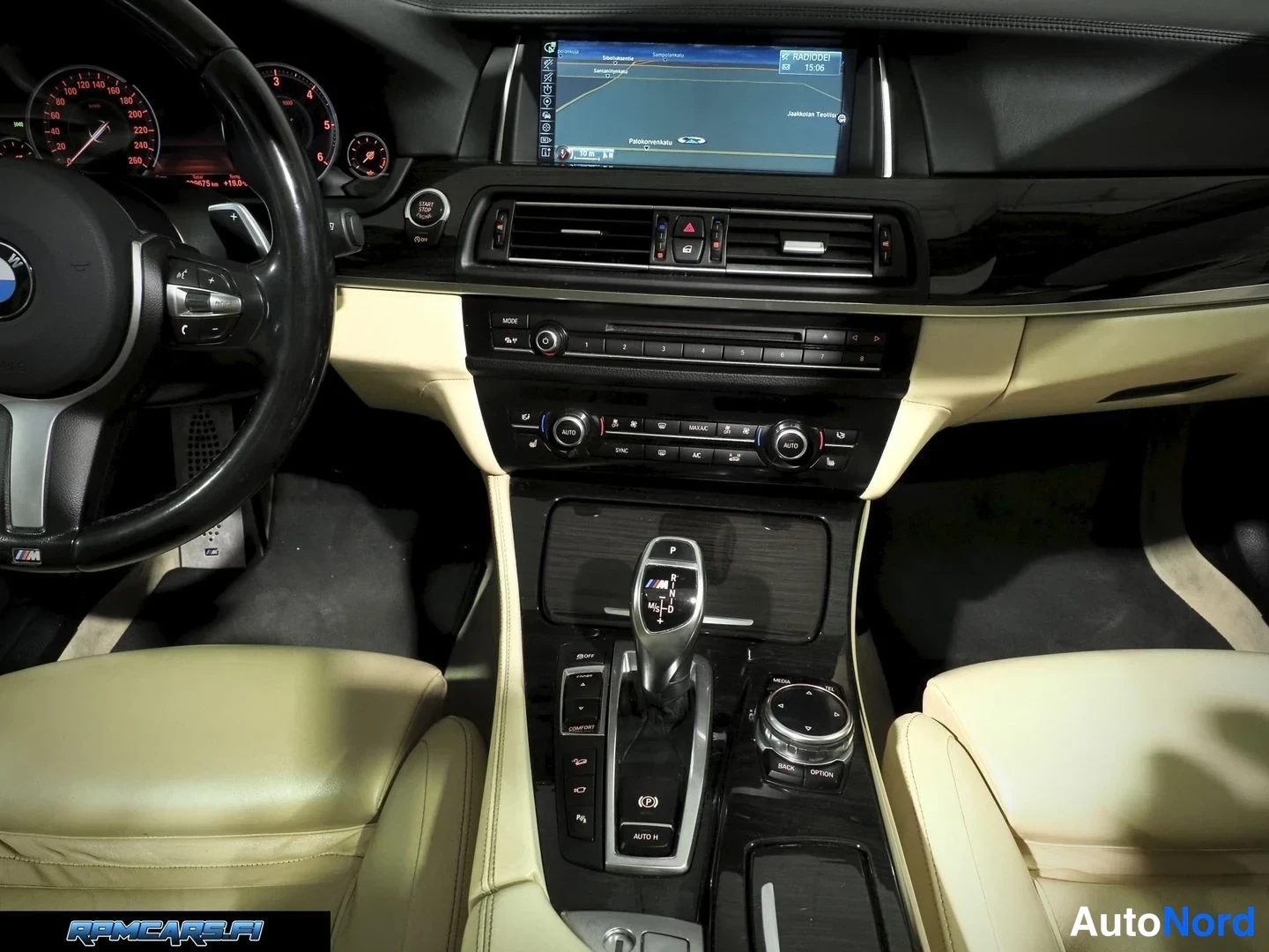 Muu: Lähikuva kojelaudasta ja keskikonsolista: BMW:n näyttö, automaattivaihteiston valitsin ja iDrive-ohjainnappi, kerma Beige-istuimet.