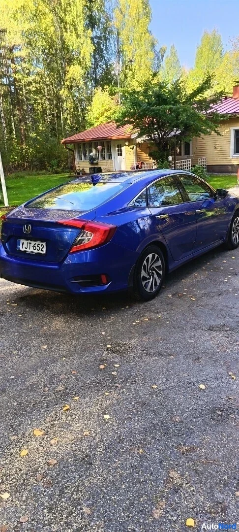Takaviisto: Sininen Honda Civic -sedan takakulmasta vasemmalta pihalla, rekisteritunnus, punakattoinen talo taustalla.