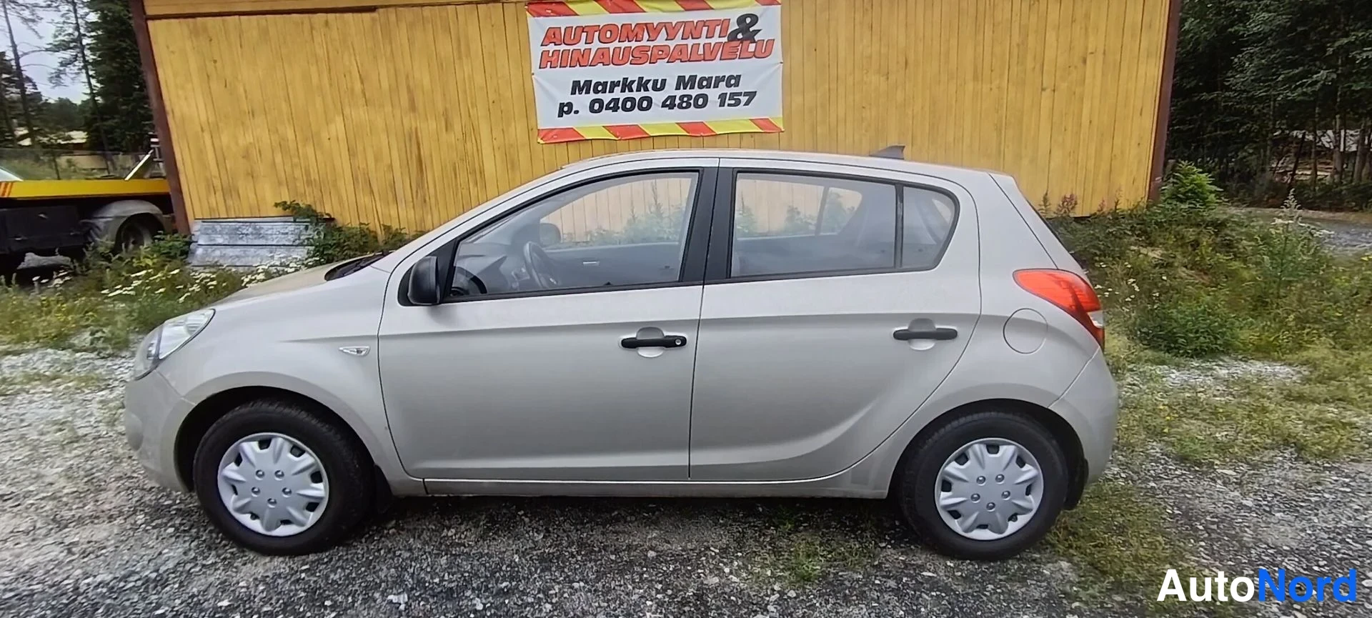 Sivulta: Hyundai i20 -viistoperä sivusta kuvattuna, vaaleanbeige kori, neljä ovea, teräsvannekset, keltaisen puurakennuksen edessä.