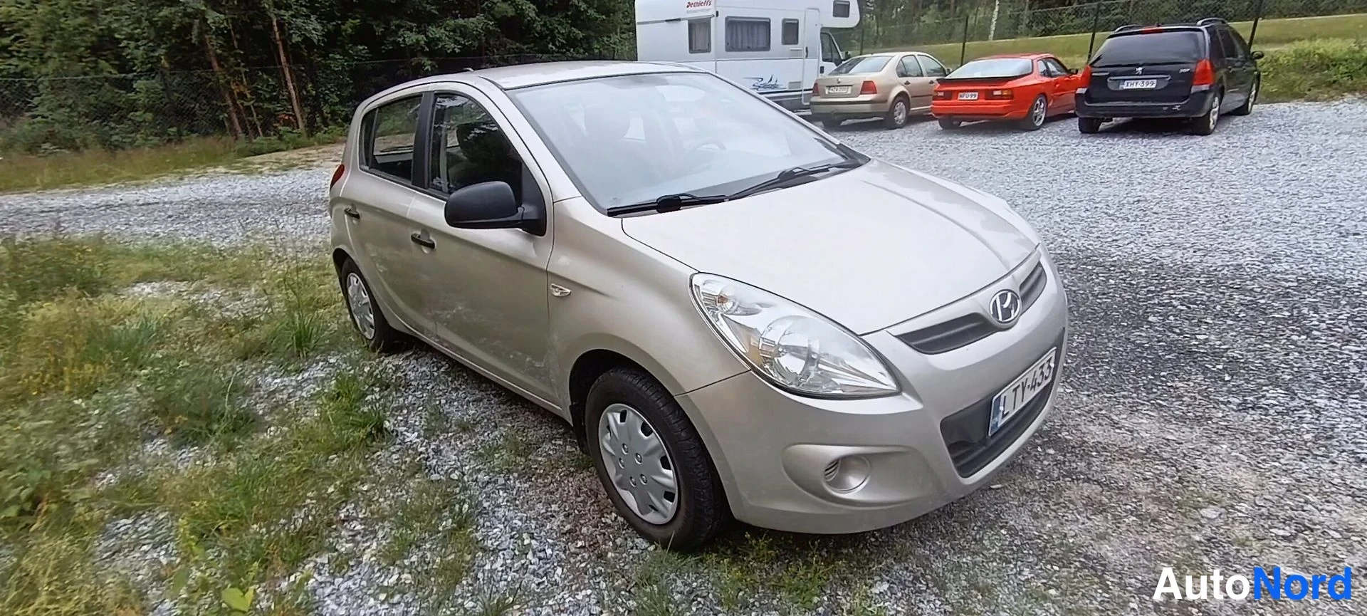 Etuviisto, nokka oikealle: Hyundai i20 etukulmasta oikealta, vaaleanbeige kori, rekisteritunnus, sora- ja nurmialusta taustalla muita autoja.