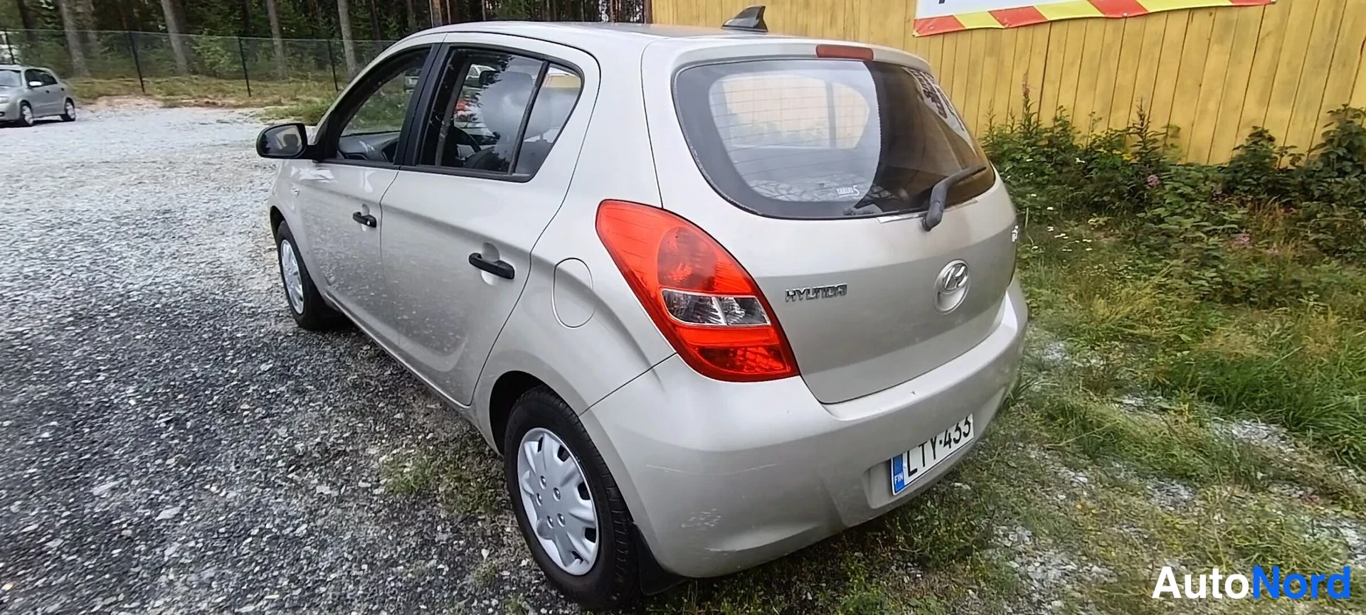 Takaviisto: Hyundai i20 -viistoperän takakulmanäkymä: beigea väriä, rekisterikilpi, takana pysäköintipaikka sorapohjaisella pihalla.