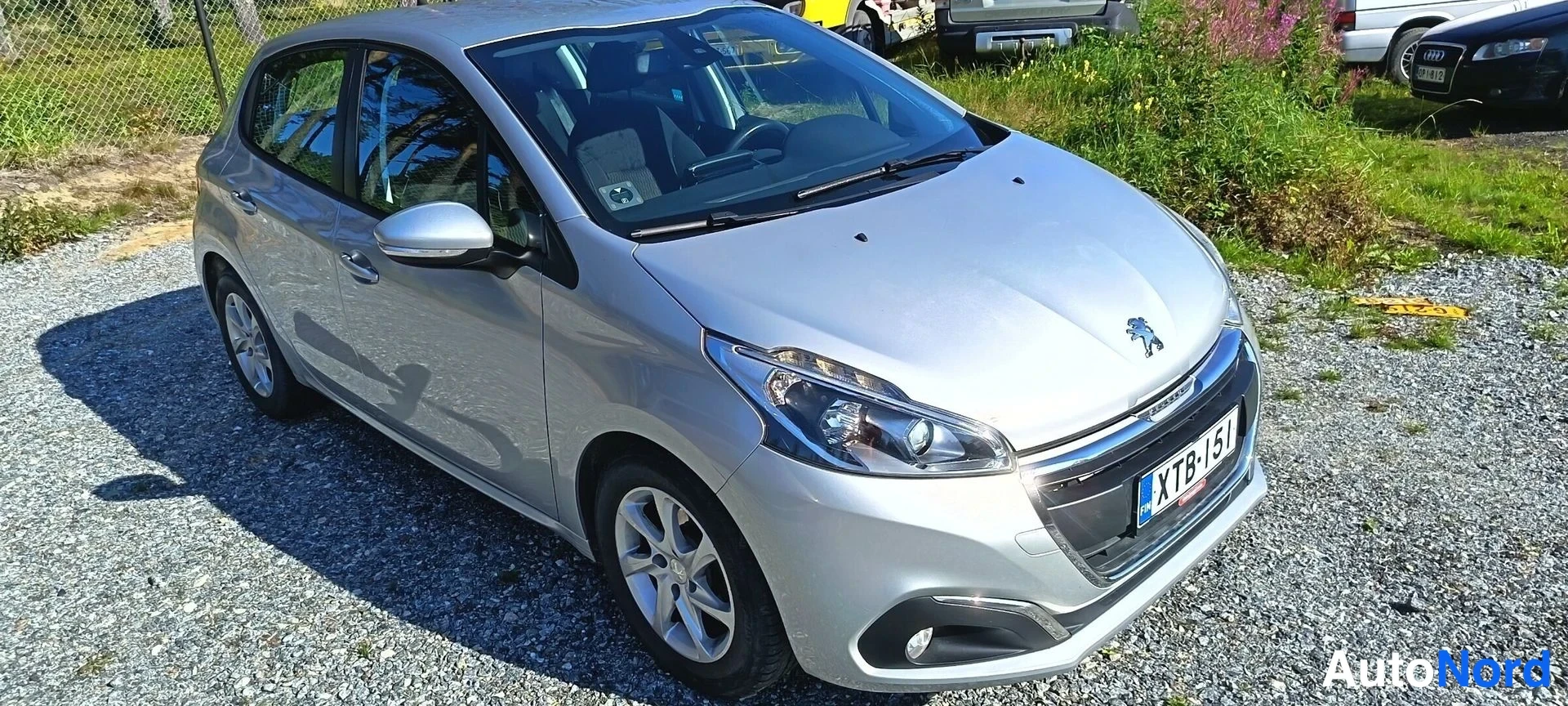 Etuviisto, nokka oikealle: Hopeanvärinen Peugeot 208 viistoperä edestä ja oikealta sivulta, rekisteritunnus.
