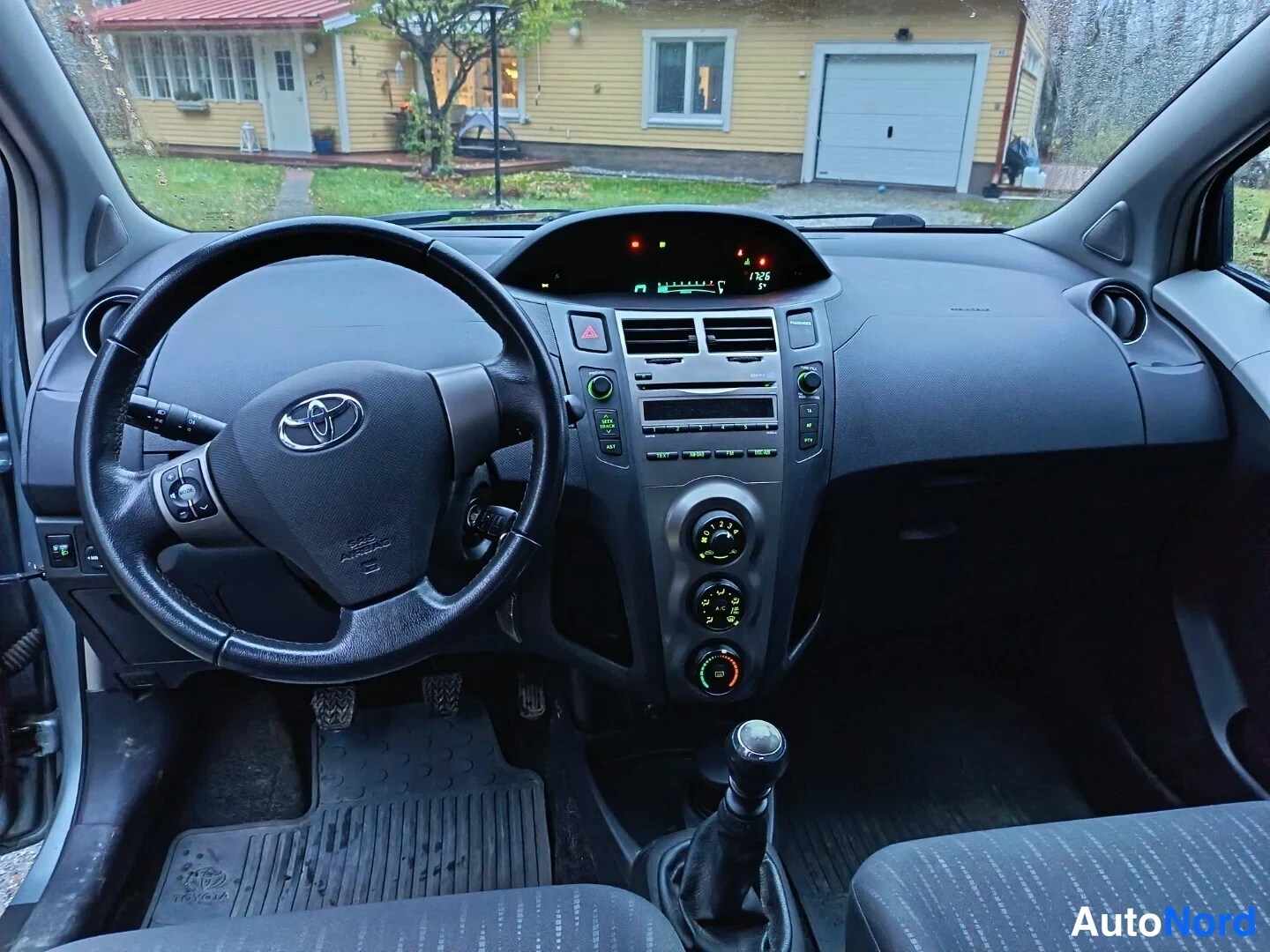 Muu: Näkymä kojelautaan kuljettajan puolelta: Toyota-merkitty ohjauspyörä, digitaalinen mittaristo, radiopaneeli, kolme säätönappia ja manuaalivaihteen vaihdinnappi.