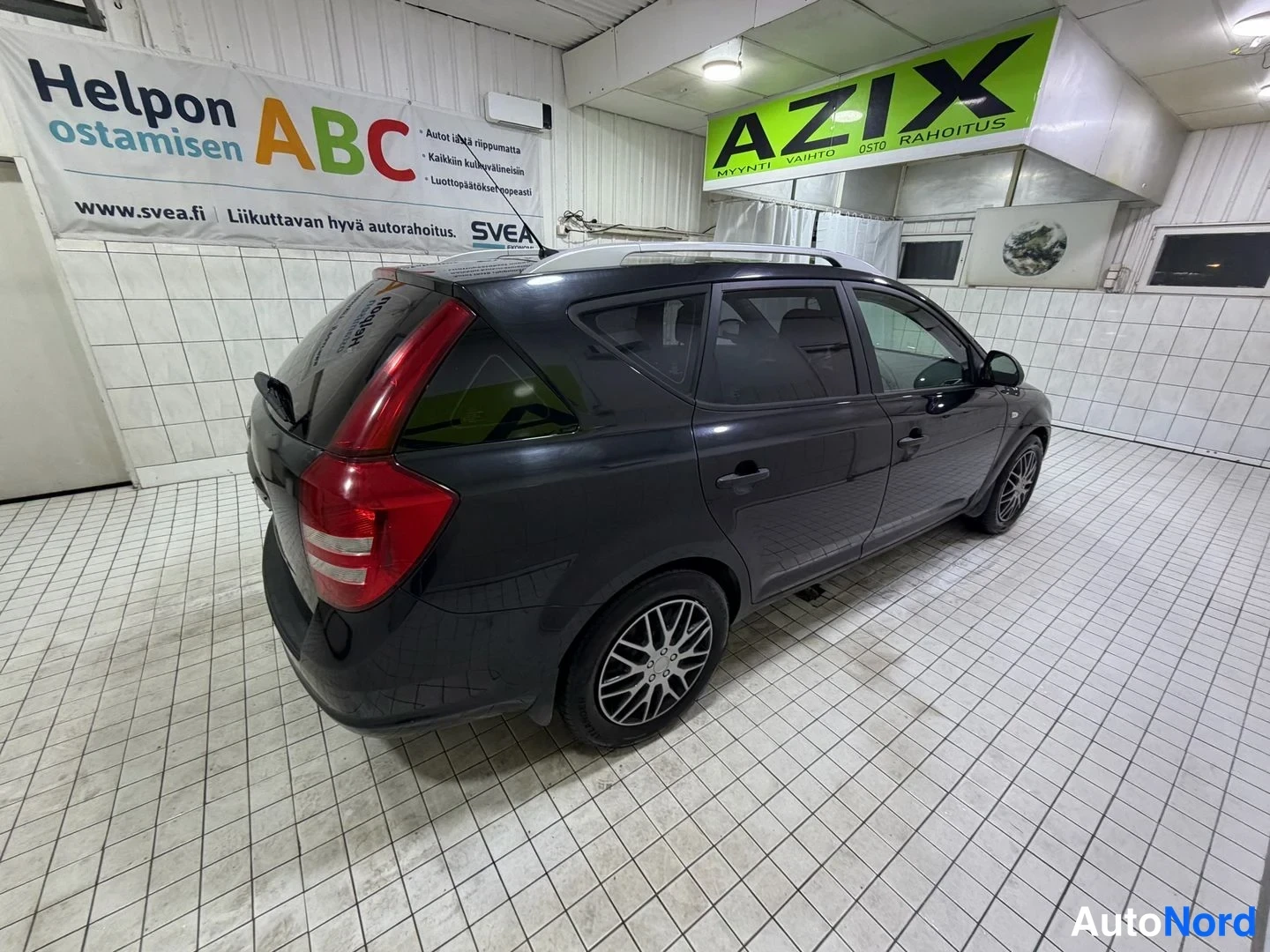 Takaviisto: Kia Ceed -farmari takaa kulmasta kuvattuna, tumma koriväri, rekisterikilpi näkyy osittain ja auton takapää kattilahuoneessa valkoisella laatoitetulla lattialla.