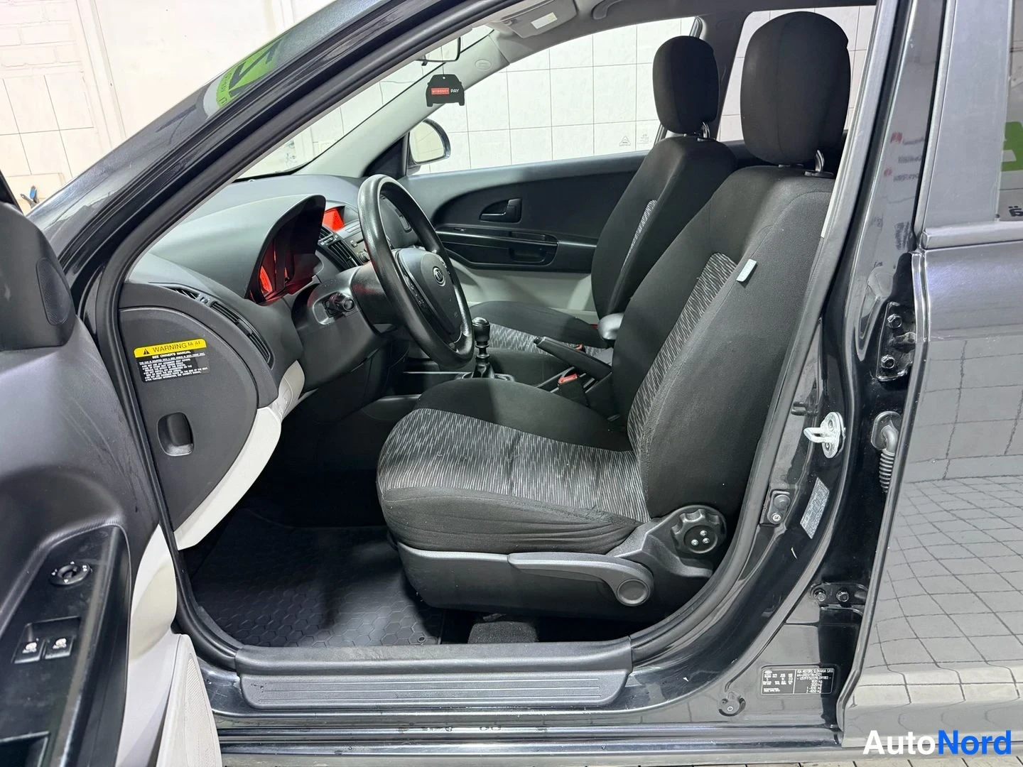 Muu: Kia Ceed -farmarin sisätilat kuljettajan oven avattua: musta kangasistuin, ohjauspyörä ja manuaalivaihteisto näkyvät.