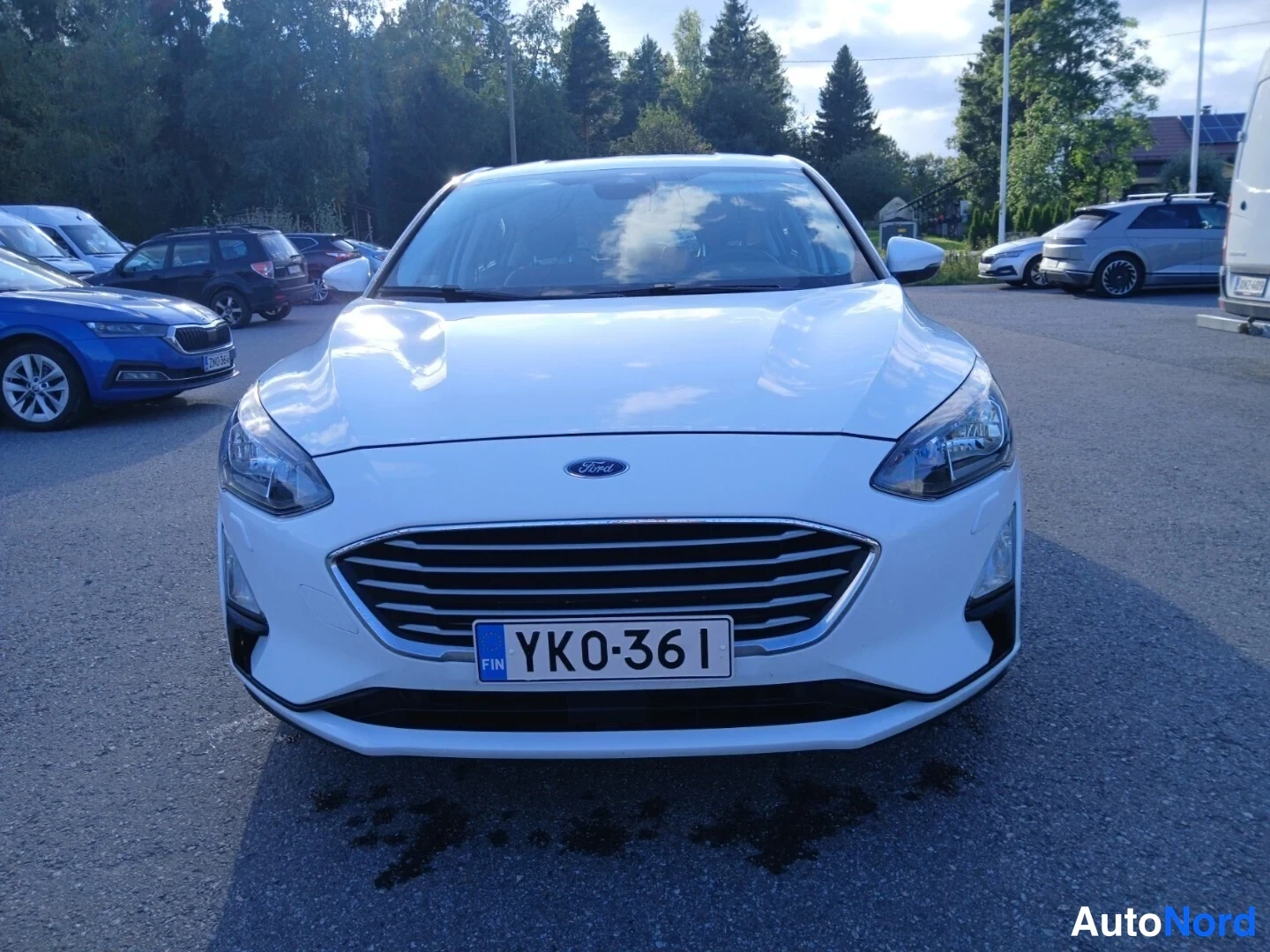 Edestä: Valkoisen Ford Focus -henkilöauton etunäkymä paikoitusalueella; Ford-merkki, jäähdytinsäleikkö, valot ja rekisterikilpi YK0-361 näkyvät.