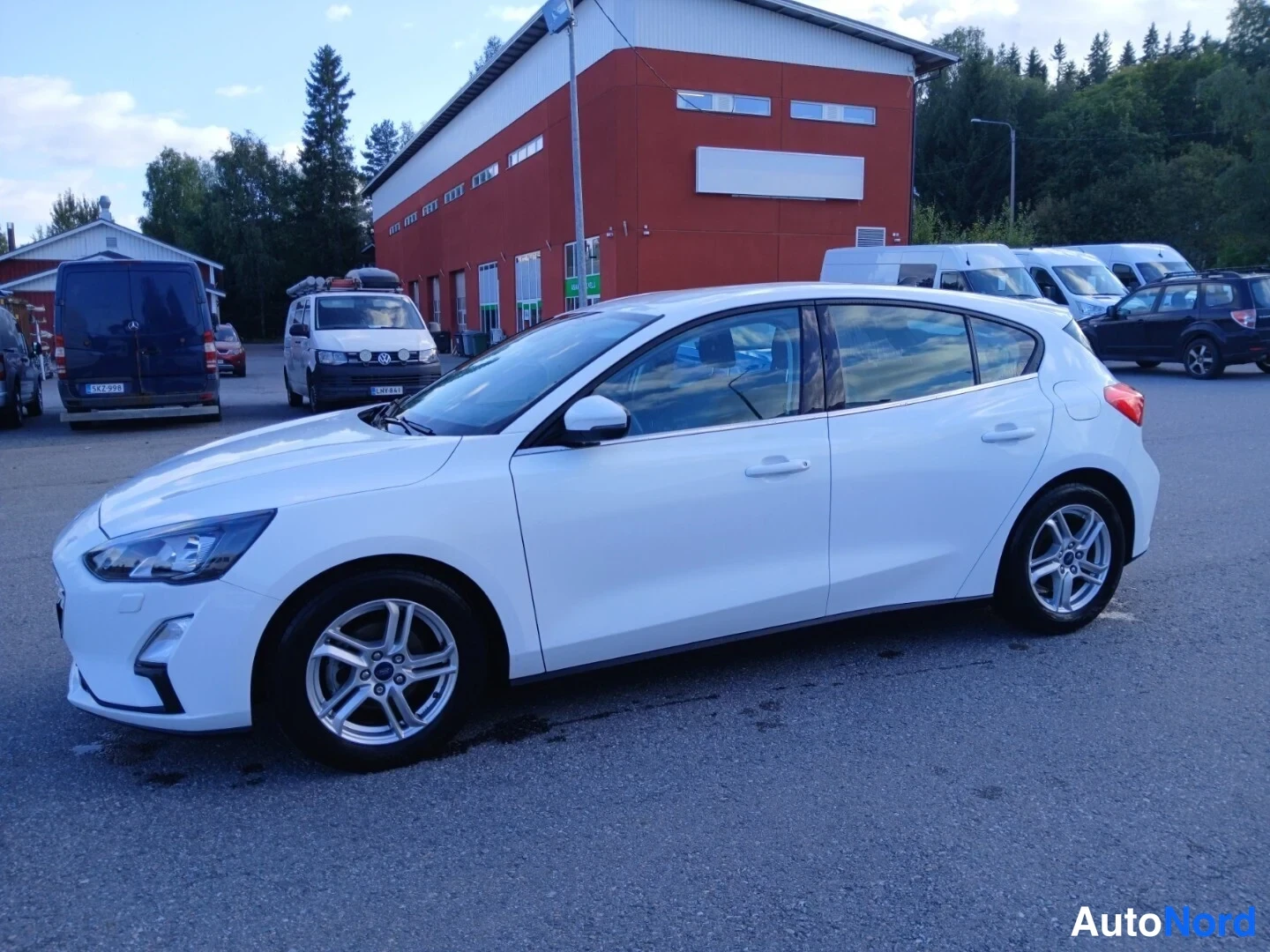 Etuviisto, nokka vasemmalle: Valkoinen Ford Focus -viistoperä sivulta edestä kuvattuna parkkipihalla, punainen rakennus taustalla.