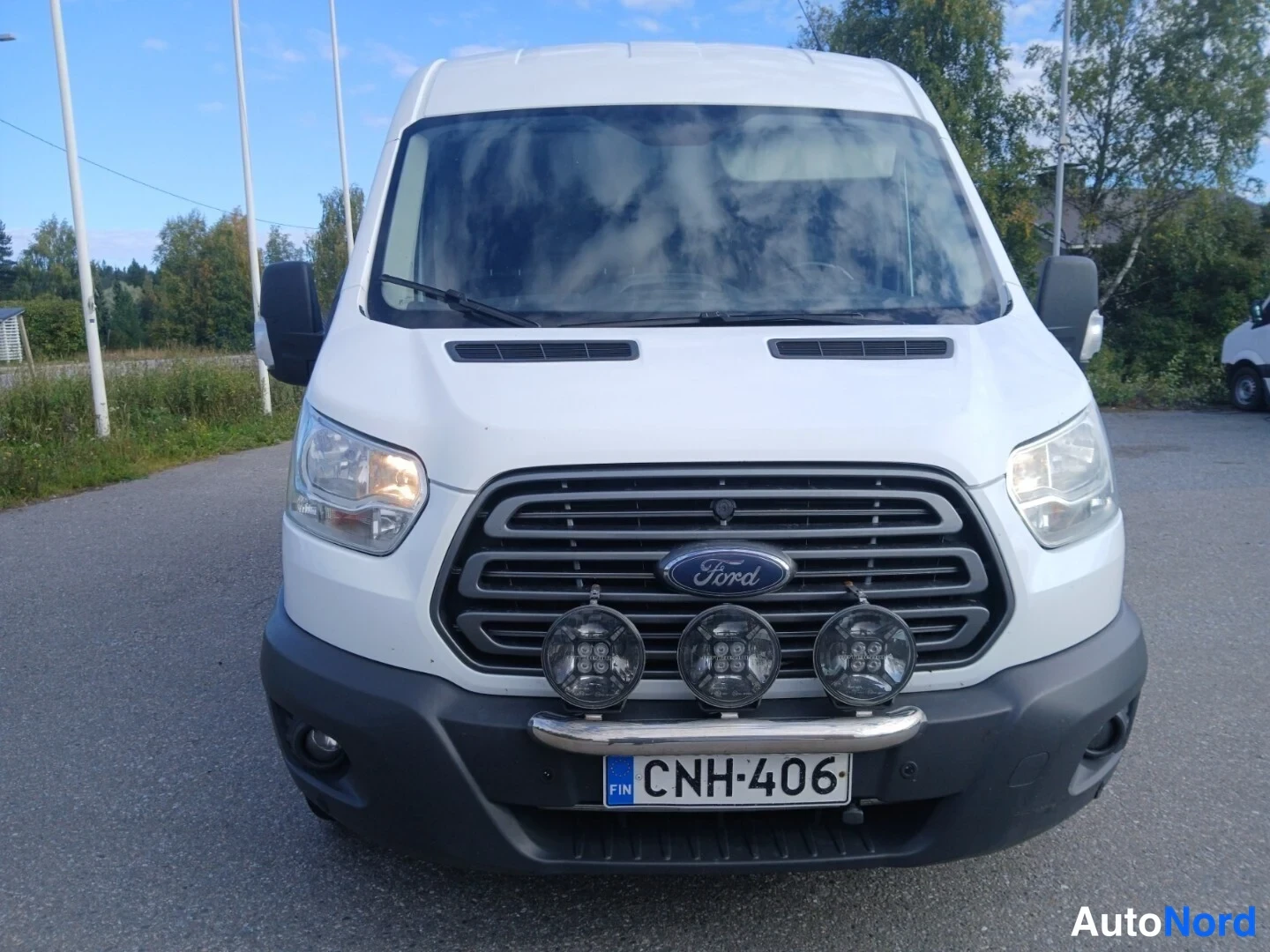 Edestä: Ford Transitin keula suoraan edestä: Ford-merkki säleikkössä, kolme lisävaloa etupuskurin kaarella, rekisterikilpi.