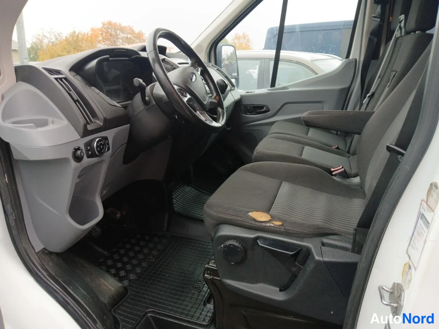 Edestä: Ford Transitin ohjaamo katsottuna etuovesta: kaksipaikkainen penkki, ratit ja tummat tekstiilimatot.