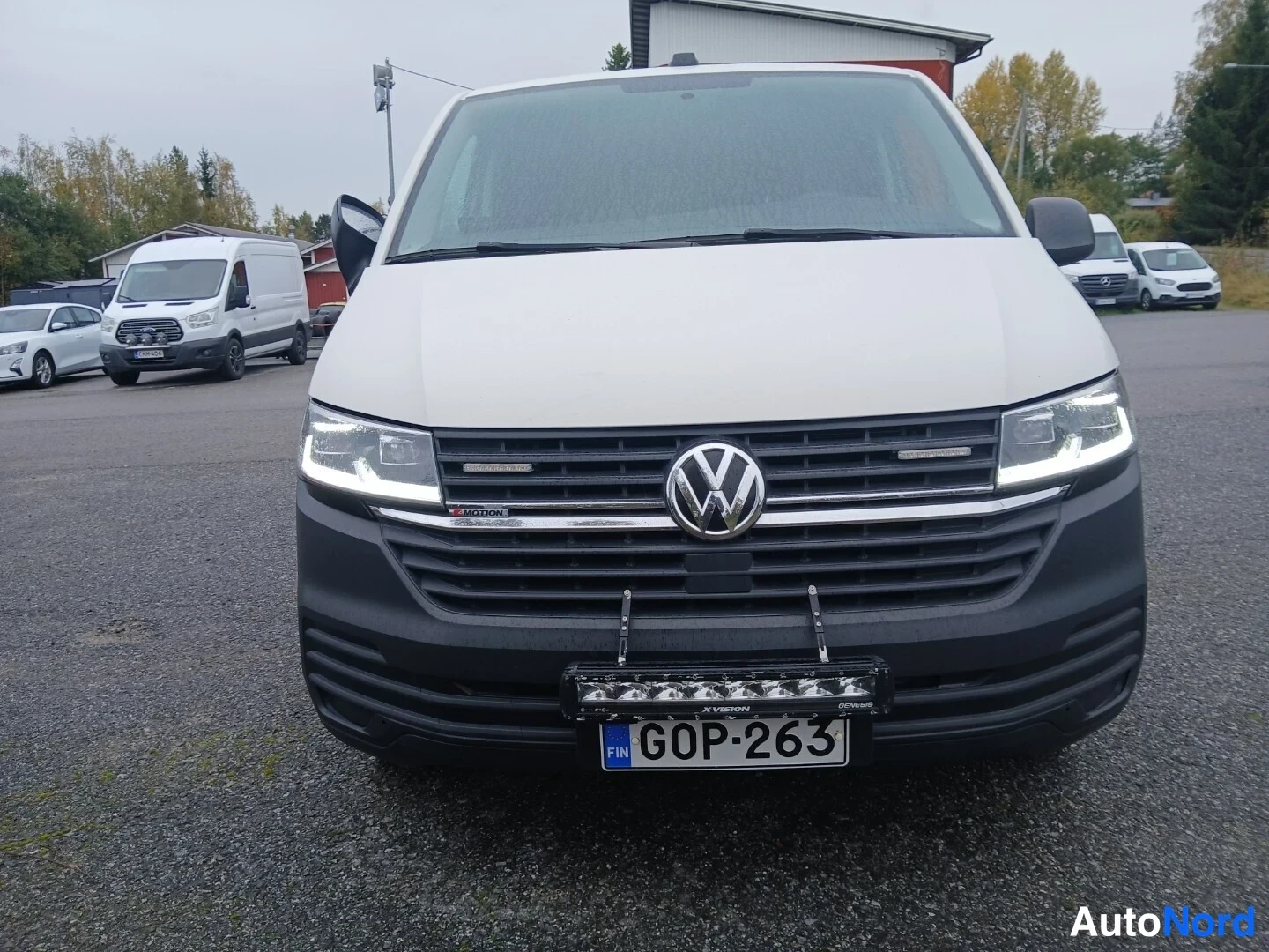 Edestä: Valkoinen pakettiauto suoraan edestä, VW-merkki ja logo kyljessä, EU-kilpi, lisävalot puskurissa.