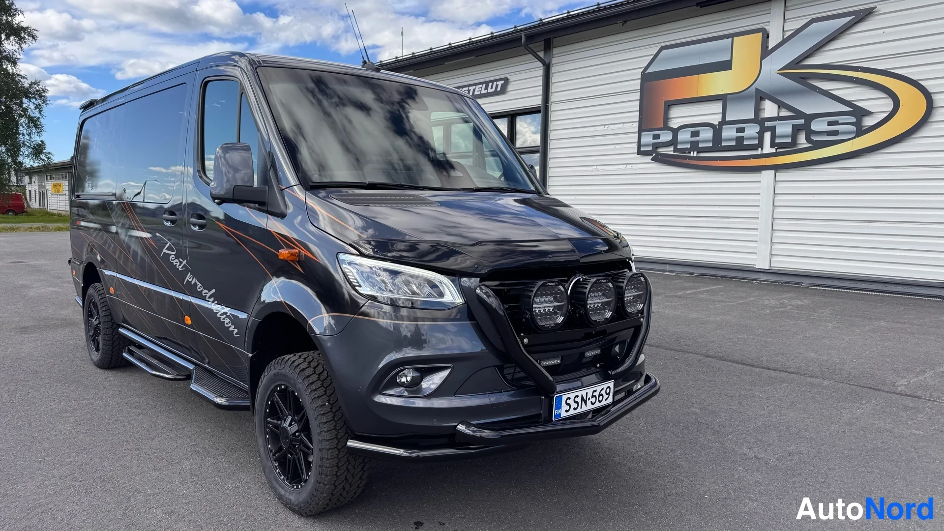 Etuviisto, nokka oikealle: Mustanharmaa Mercedes-Benz Sprinter -pakettiauto etuokolikuvassa, tummat kevytmetallivanteet ja lisävalot etupuskurin kaarella.