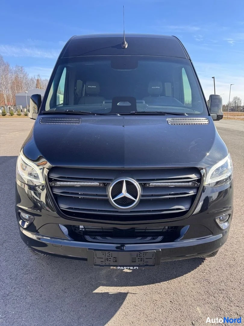 Edestä: Mustan Mercedes-Benz Sprinter -pakettiauton etuosa suoraan edestä: tähtimerkki etusäleikössä, LED-ajovalot, tummat ikkunaluukkutunnustimet sisätilassa ja tyhjä