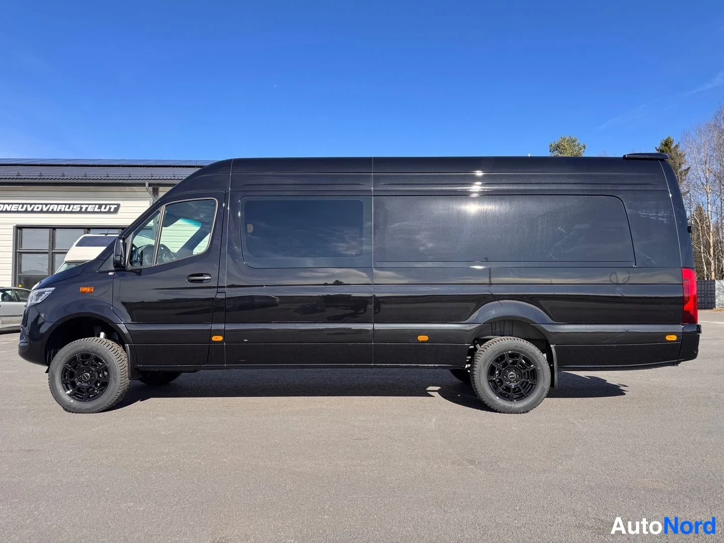 Sivulta: Mustan Mercedes-Benz Sprinter -pakettiauton sivuprofiili vasemmalta: pitkä korirakenne, tummat ikkunat, mustat kevytmetallivanteet ja takapuskurin punainen kivi