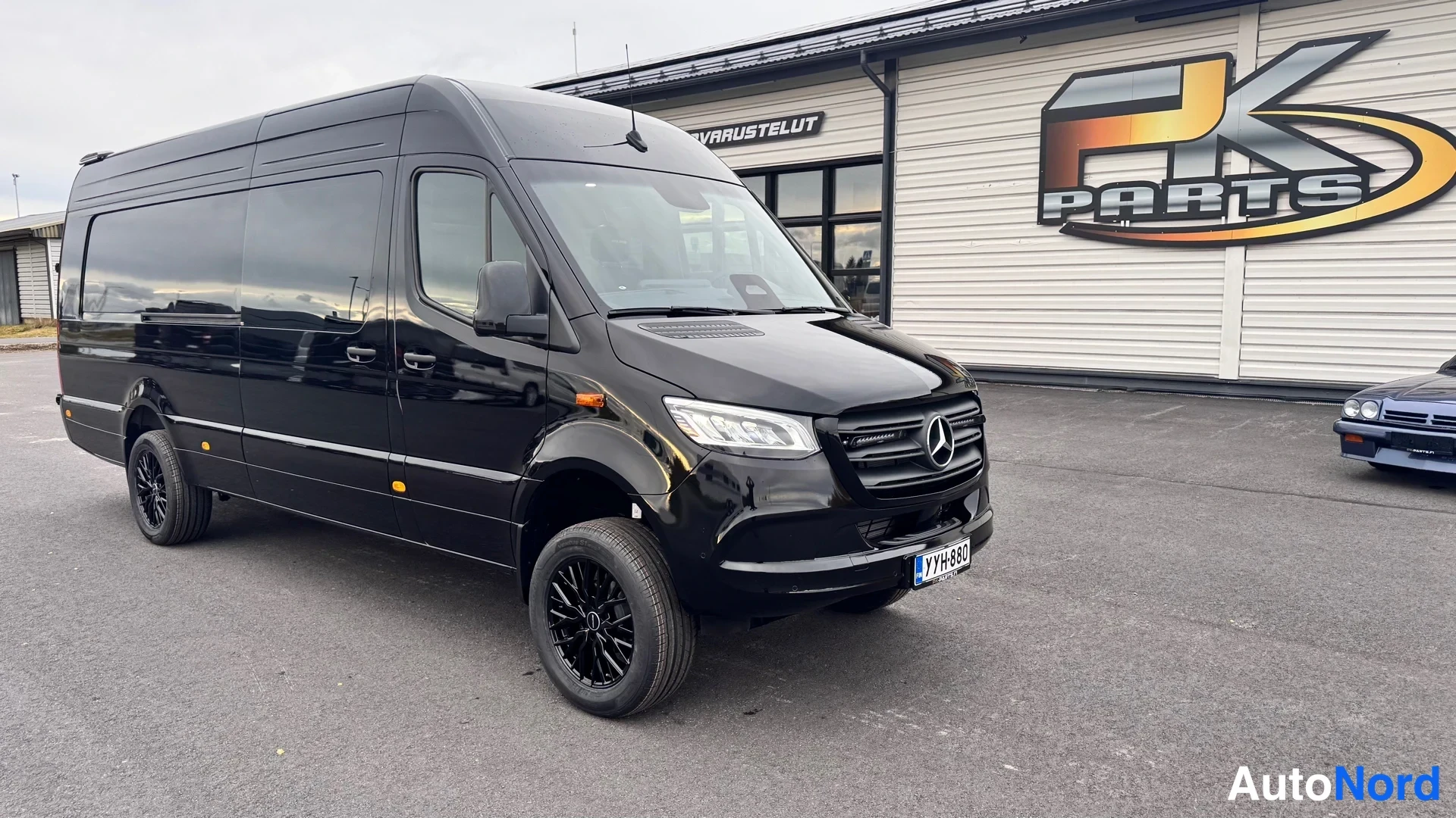 Etuviisto, nokka oikealle: Musta Mercedes-Benz Sprinter -pakettiauto etuokolikuvassa, kevytmetallivanteet ja etulokasuojat.