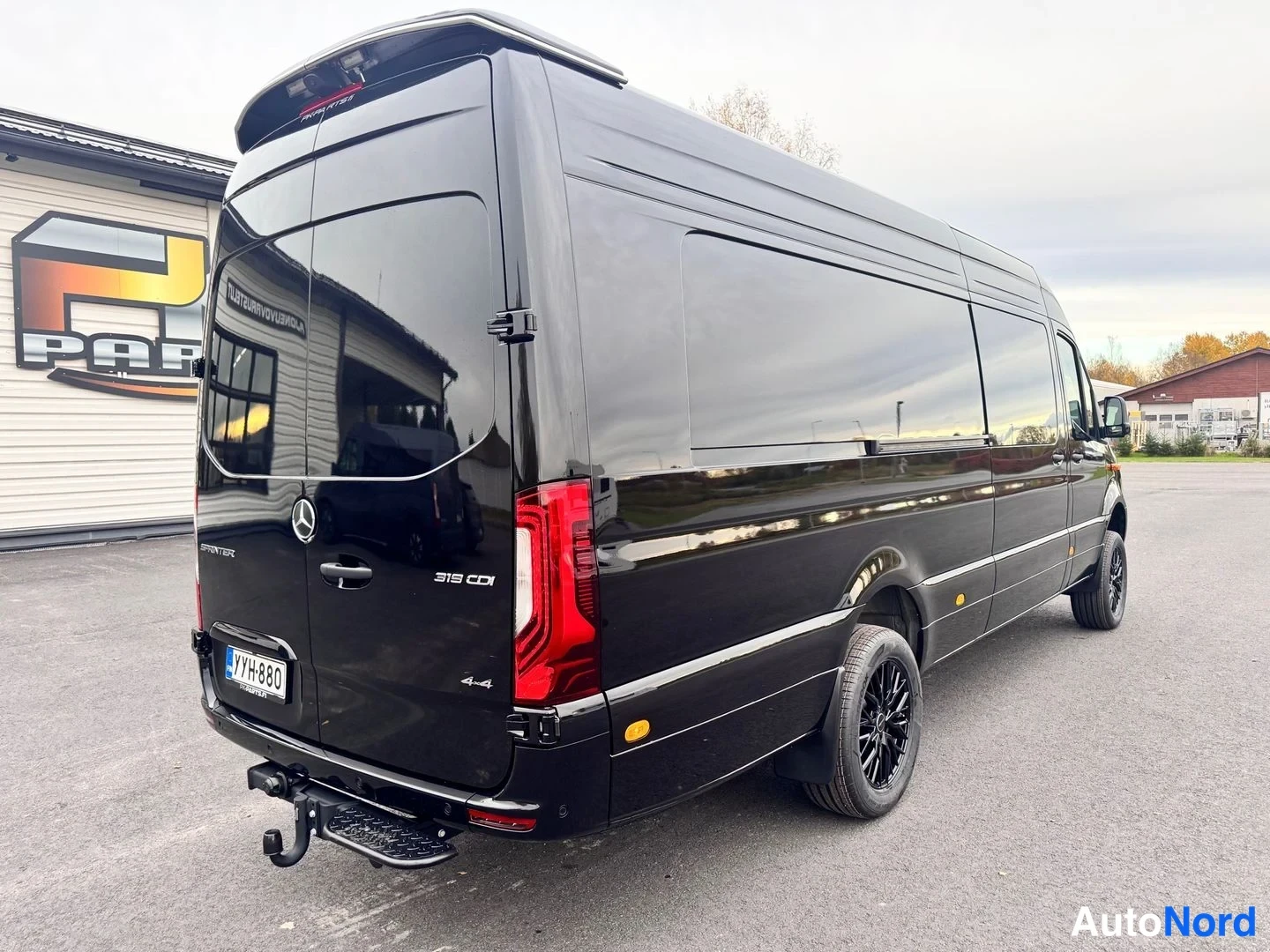 Takaviisto: Musta Mercedes-Benz Sprinter -pakettiauto kuvattu takaa viistosti; takamoottoritilan kannessa Mercedes-tähti, takapuskurissa kytkysilmukka, rekisteritunnus.