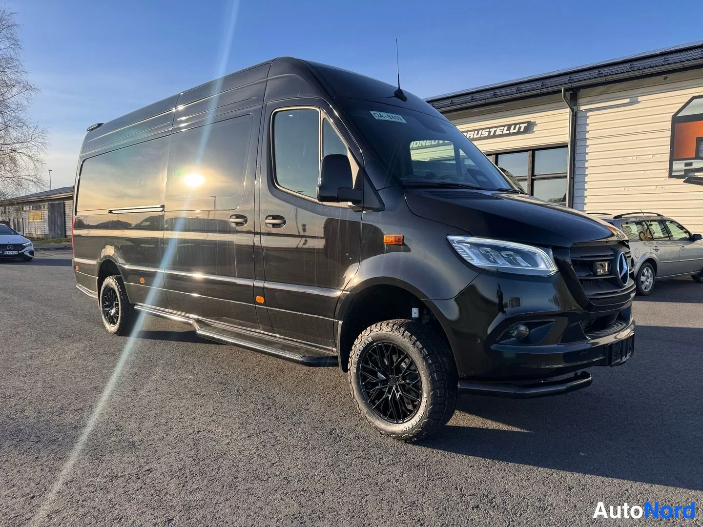 Etuviisto, nokka oikealle: Musta Mercedes-Benz Sprinter -pakettiauto etuviistosta oikealta; tummat kevytmetallivanteet, sumsuoja ja lisävalo etupuskurissa.